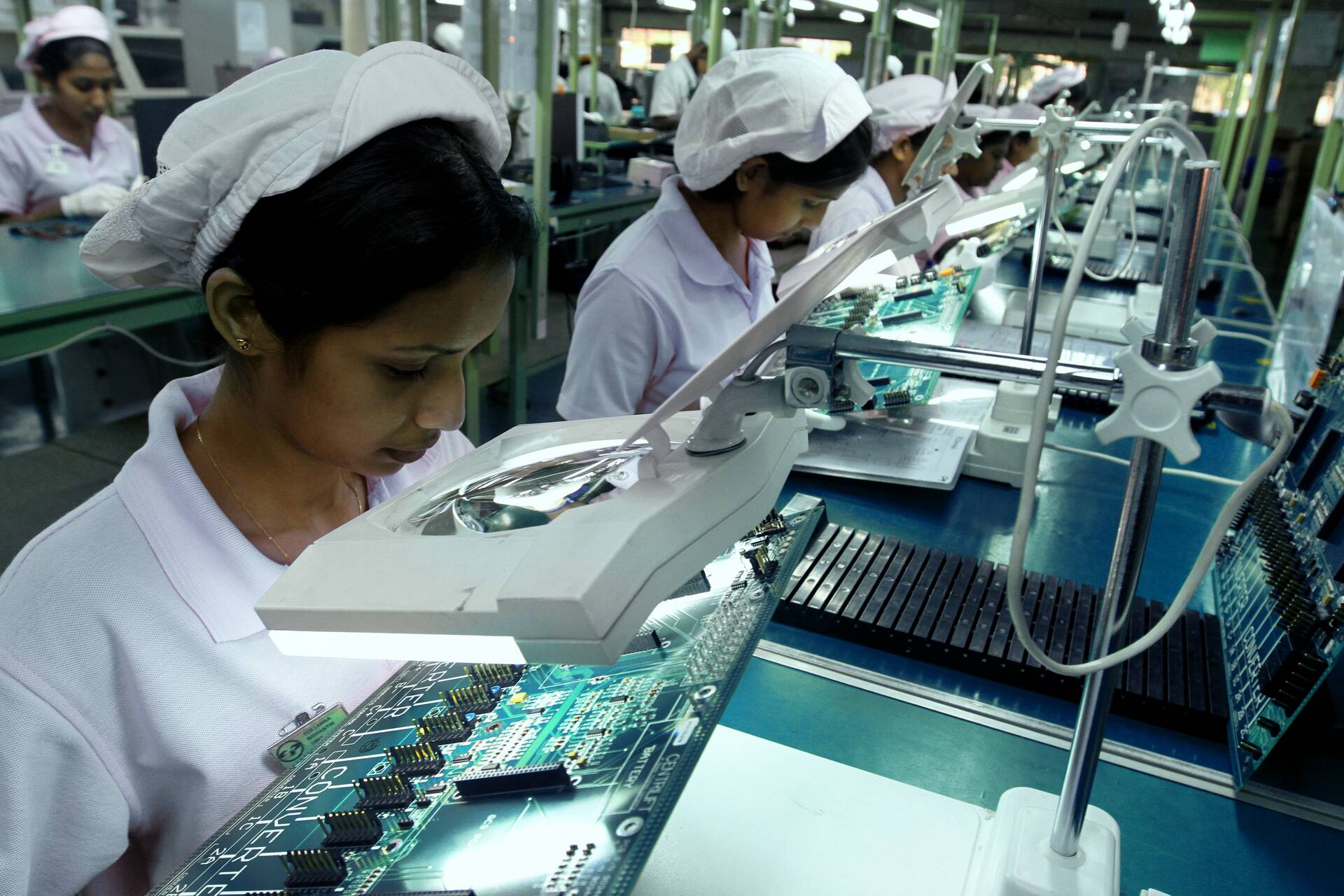 female workers with disposable head caps inspecting PCB production lines through magnifying lenses
